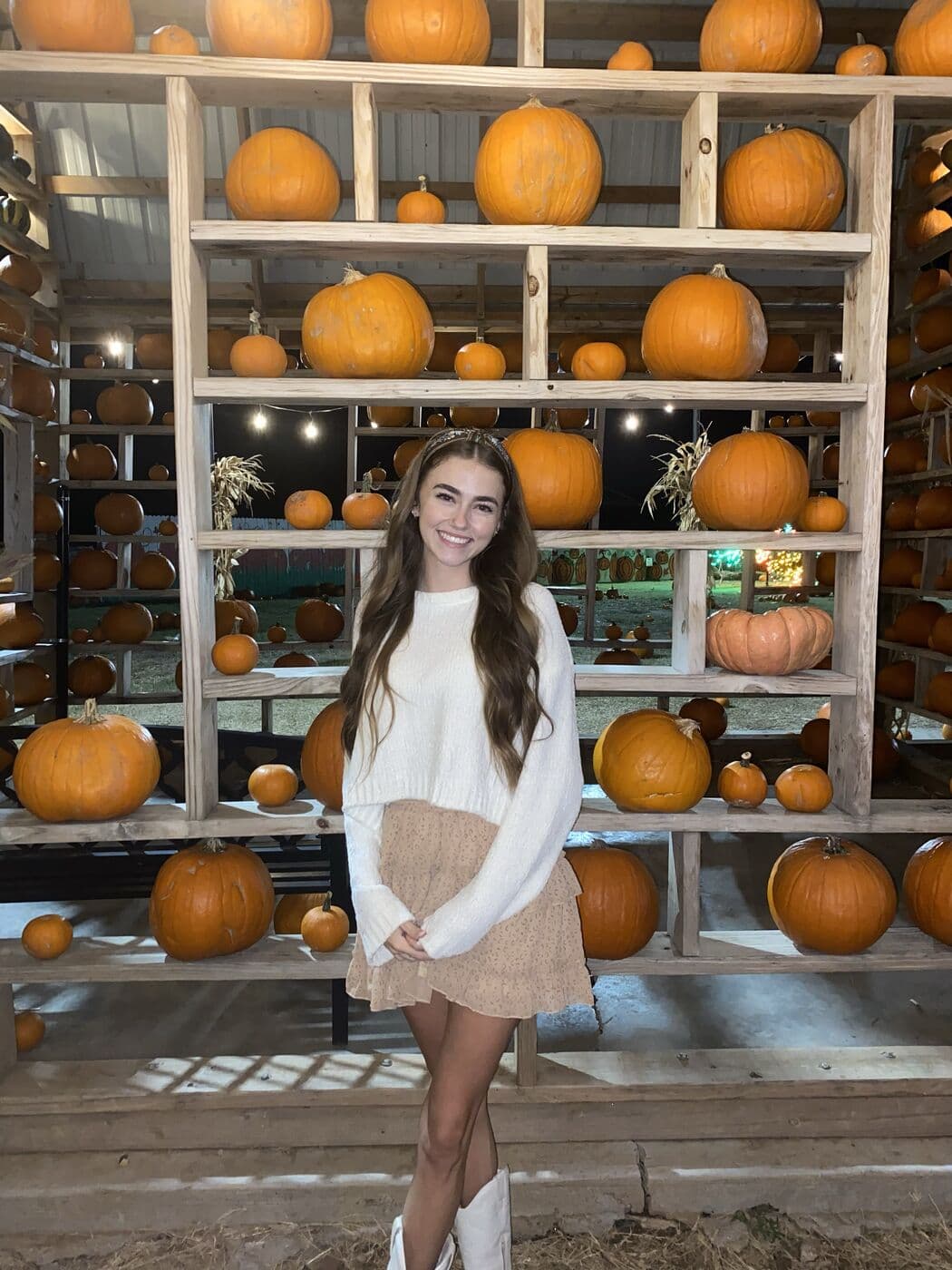 Elise standing in front of the pumpkin shelves in a white sweater and boots